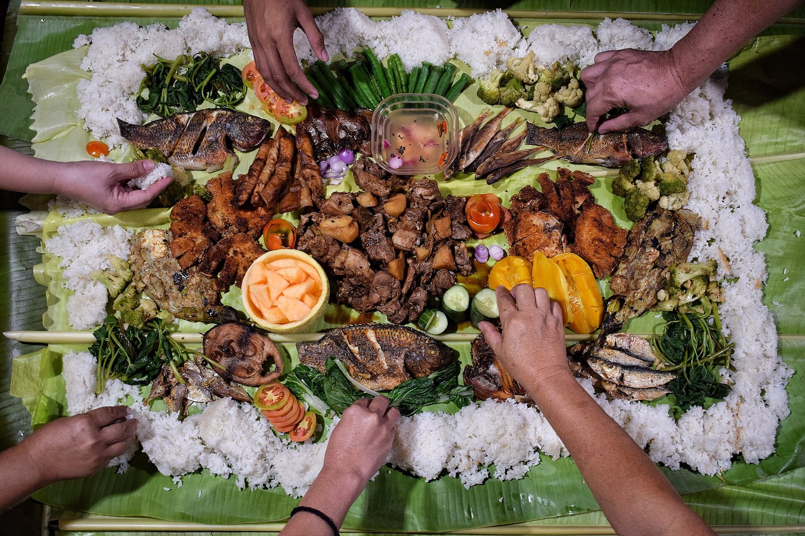 Hands reaching in to share a kamayan feast laid out on banana leaves