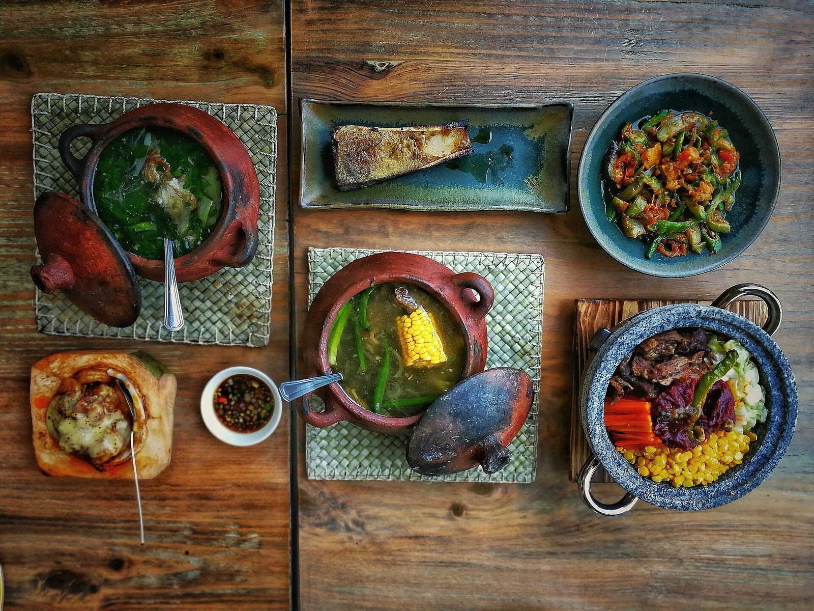 A rustic wooden table set with Filipino dishes in clay pots and bowls, seen from above